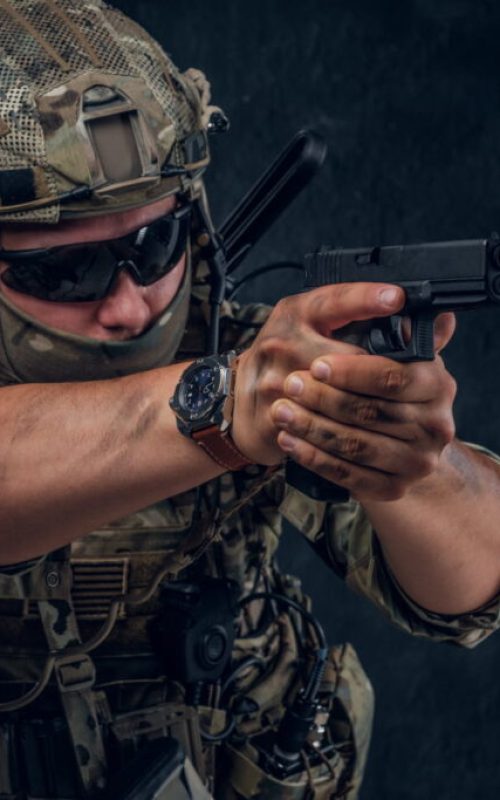 Serious brave soldier in military uniform and sunglasses is aiming with his gun.